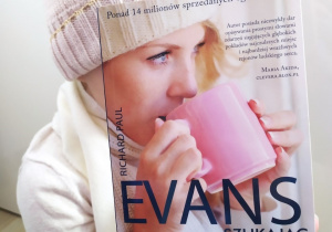 Praca Kingi Marciniak - Zdjęcie przedstawia młodą dziewczynę ubraną w ciepły sweter, czapkę i szalik w kolorze białym. Twarz zastąpiono okładką książki „Szukając Noel” Richarda Paula Evansa.